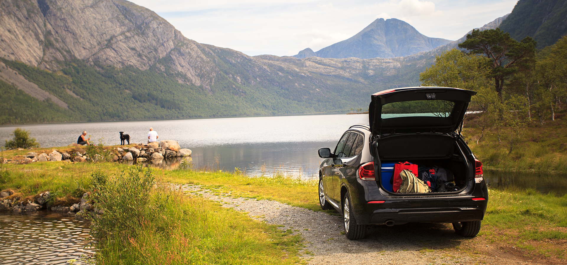 A small SUV parked on a gravel path near a lake with the trunk open as a couple and dog sit on the bank nearby.