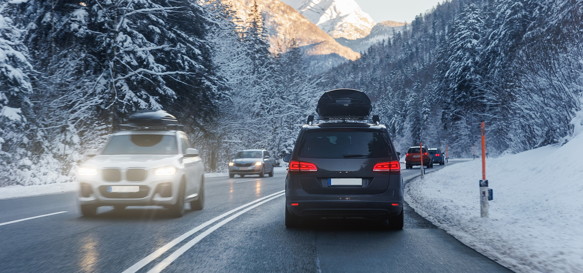 Several vehicles driving on a wet roadway, surrounded by snowy wilderness, with mountains in the background.